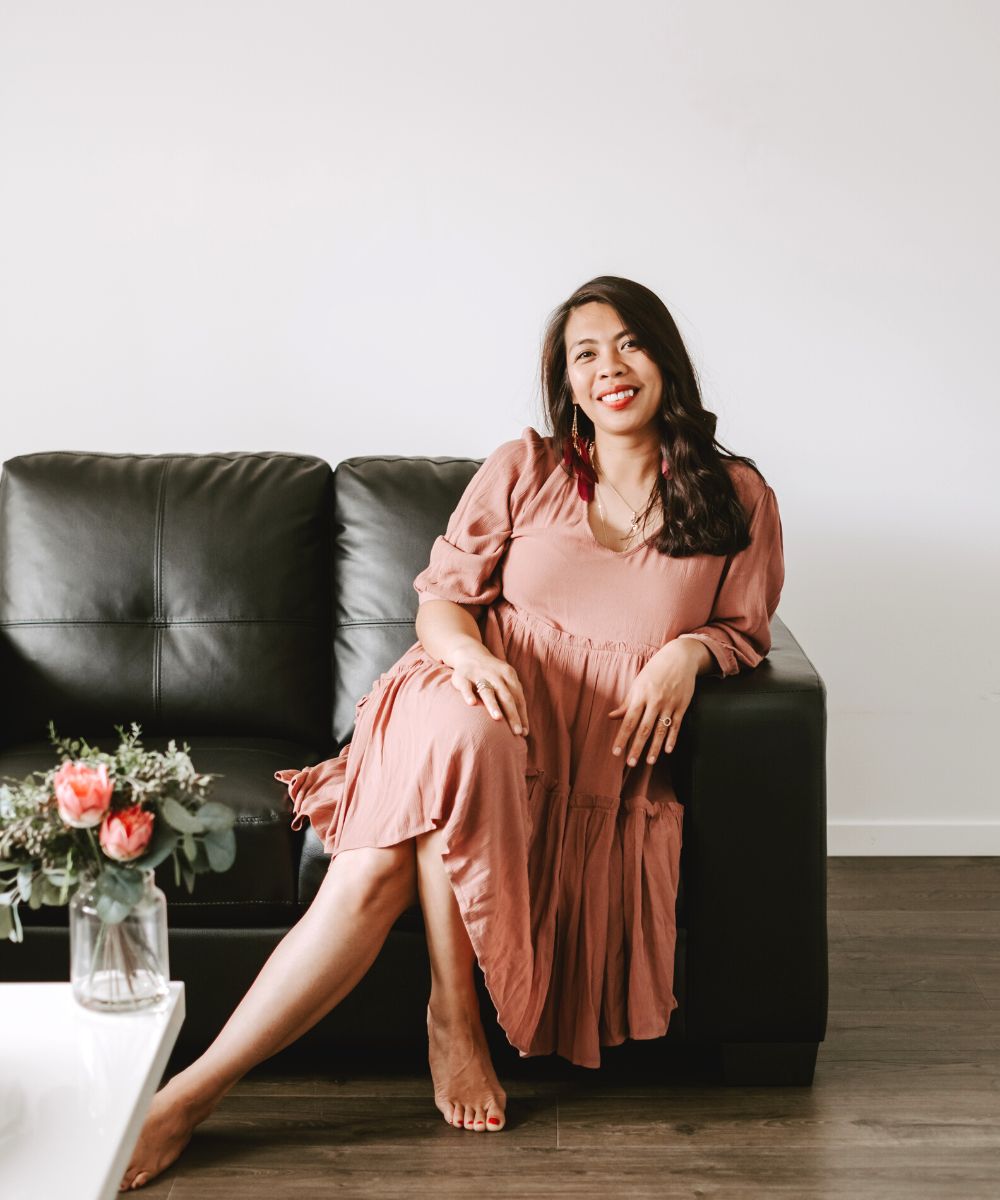 Michelle is smiling wearing a ping dress and a crystal necklace. She is sitting on a black leather couch. In front of her is a white coffee table with a vase of pink roses in the corner.
