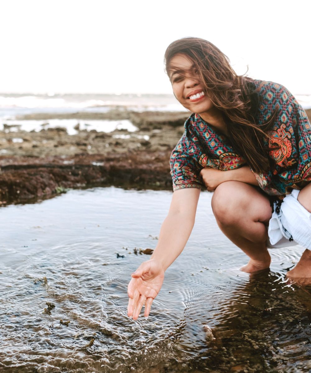 Michelle is at the beach, splashing water from a pool of rocks. She is wearing a white dress and has a colorful scarf around her shoulders. The wind is blowing her hair on one side and she is happy.
