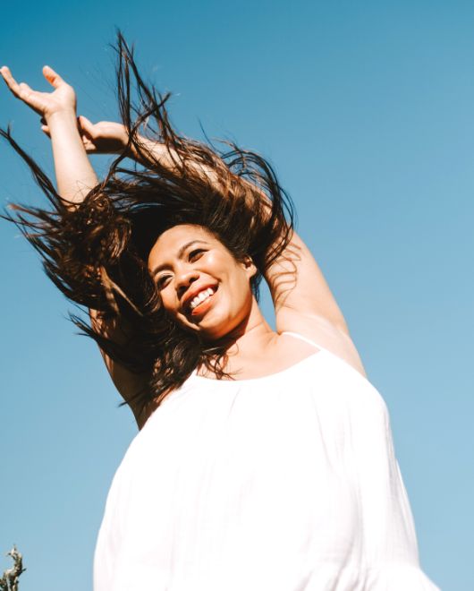 Michelle is wearing a white dress and is smiling. the wind is blowing her hair and she has her arms up to the blue sky.